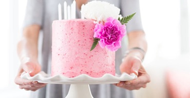 photo of a mdeium sized strawberry cake on top of a white pedestal. decorated with a pink and white flower. held at arms length by someone presenting it to the camera