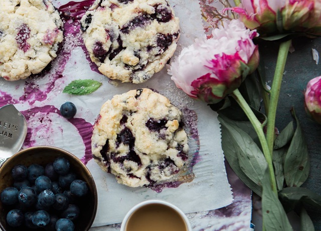 top down photo of sliced open blueberry muffins with blue berries in a bowl on the side and 2 large flowers with stems on the side.